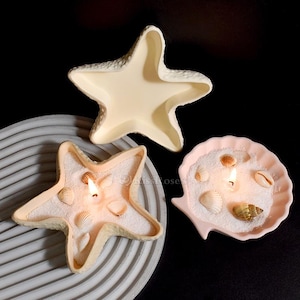 May include: Three decorative candles in the shape of a starfish and a seashell. The starfish candles are filled with white sand and seashells, with a lit wick. The seashell candle is pink and also contains sand, seashells, and a lit wick.