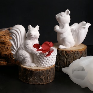 Puede incluir: Figuras blancas de ardillas, una con una cesta con flores rojas, la otra con una maceta en forma de tulipán. Ambas sobre rodajas de madera. Figuras decorativas para el hogar.
