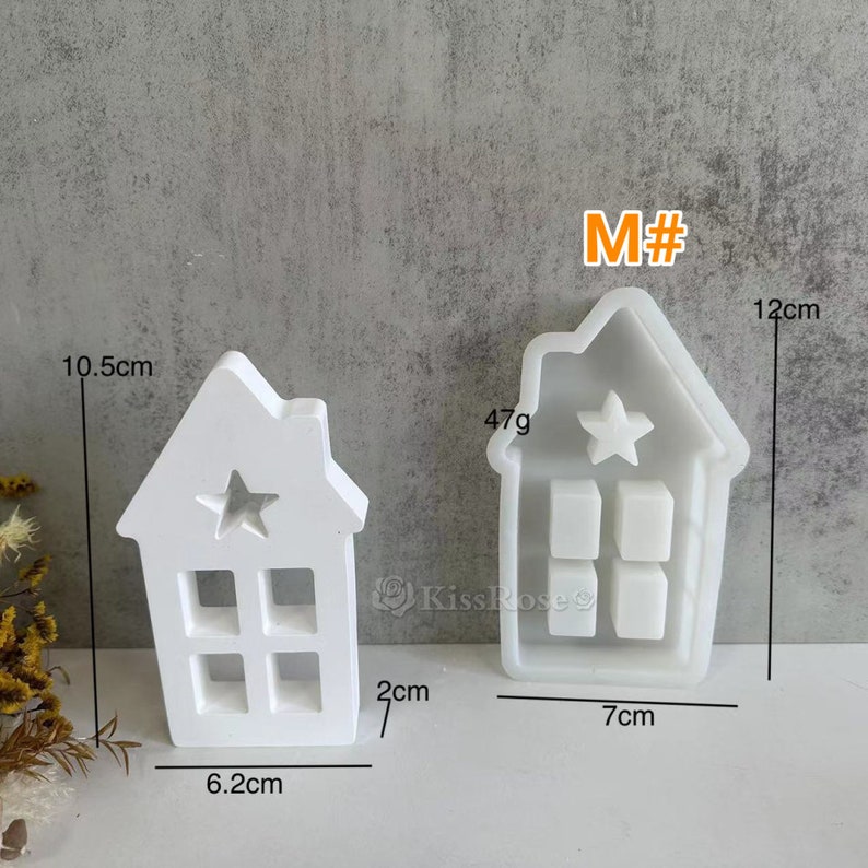 Może przedstawiać: Dwa białe plastikowe formy w kształcie dom&oacute;w z detalami gwiazd i okien. Większa forma ma 12 cm wysokości i 7 cm szerokości. Mniejsza forma ma 10,5 cm wysokości i 6,2 cm szerokości. Formy są oznaczone "M#" i "47g".