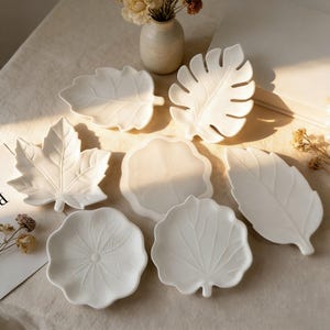 May include: A collection of seven white ceramic dishes shaped like various leaves and plants, including a maple leaf, a monstera leaf, and rounded foliage, arranged on a light-colored surface with a small ceramic vase and dried flowers.