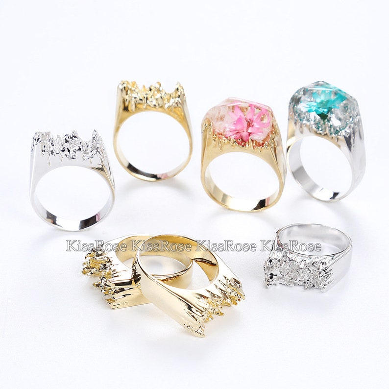 May include: Five silver and gold rings with a jagged edge design. Some rings have a clear resin top with dried flowers inside. The rings are on a white surface.