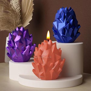 May include: Three geometrically shaped candles, one lit, in shades of blue, purple, and orange. The candles are arranged on a tiered white platform.