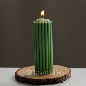 May include: A green pillar candle with a ribbed design is lit and sitting on a wooden slice. The candle is on a white surface.