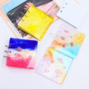 Puede incluir: Cuatro cubiertas de carpeta de resina diferentes. Una es amarilla con purpurina, otra es azul y rosa con purpurina, y dos son multicolores con flores blancas y rosas.