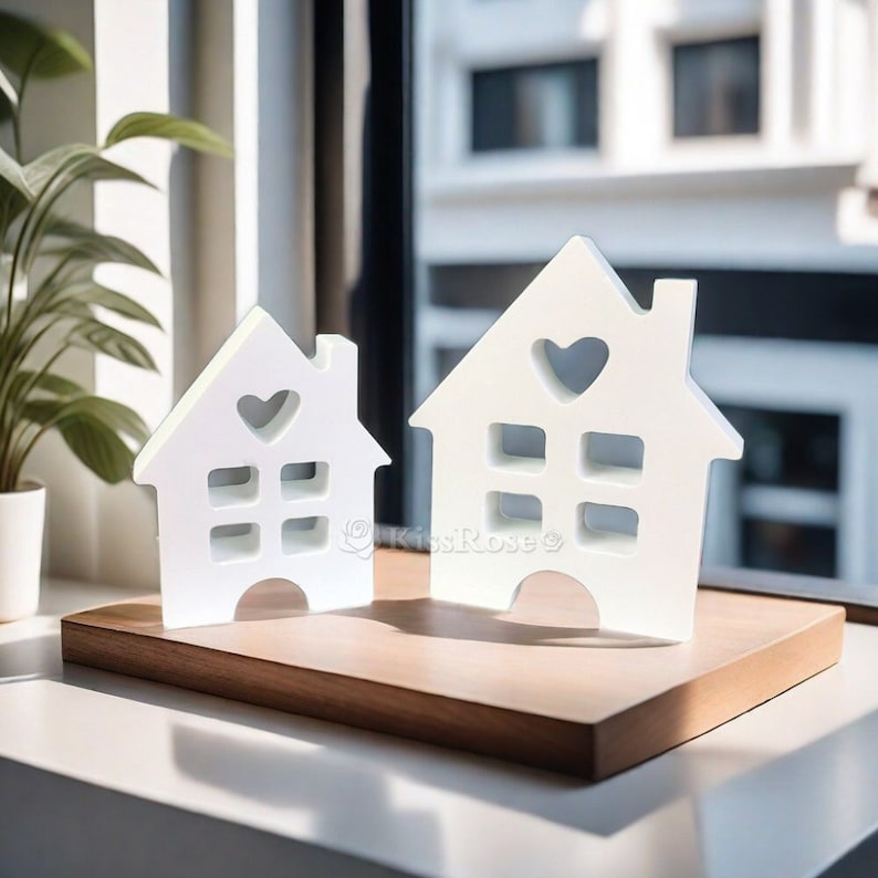 May include: Two white wooden house shaped ornaments with heart cutouts and window details. The ornaments are sitting on a wooden base.