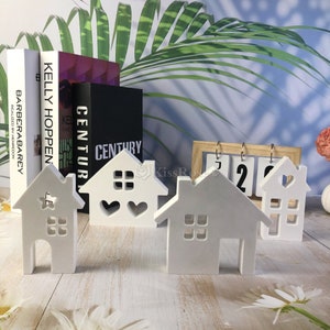 Pu&ograve; includere: Quattro case di legno bianche con finestre e cuori ritagliati. Le case sono disposte su una superficie di legno con un calendario sullo sfondo.