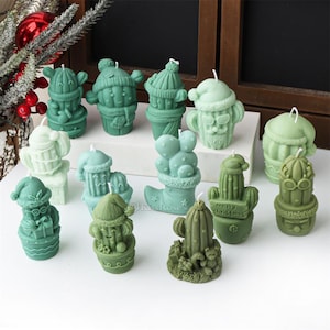 Puede incluir: Surtido de velas con forma de cactus navideño de color verde y blanco. Las velas presentan varios diseños, incluyendo gorros de Papá Noel, botas y cactus con las palabras "Feliz Navidad".
