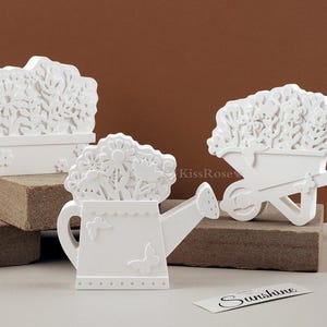 Puede incluir: Tres figuritas decorativas blancas con temática de jardín. La primera figurita es una maceta con un diseño floral. La segunda figurita es una regadera con un diseño floral. La tercera figurita es una carretilla con un diseño floral. Las figuritas están sobre una superficie de madera con un letrero que dice "Sunshine".