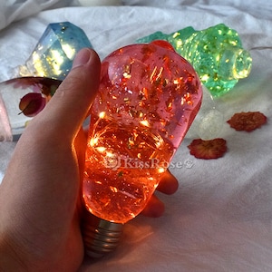 May include: A clear glass light bulb filled with red resin and gold flakes. The bulb has a string of warm white LED lights inside. The bulb is being held by a hand.