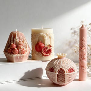 May include: A collection of decorative candles in various shapes and designs. Includes a pink geometric candle with a castle motif, a cream-colored pillar candle with pomegranate details, a pink pomegranate-shaped candle, and a pink pillar candle with fruit carvings.