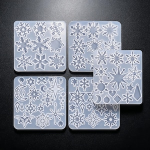 May include: Four translucent silicone molds featuring various shapes including snowflakes, stars, moons, and geometric pendants. Each square mold contains multiple recessed cavities designed for casting small decorative items with intricate cut-out details.