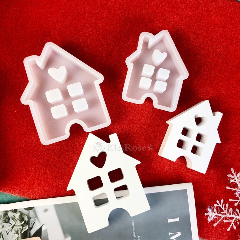 May include: Four white silicone molds shaped like houses with heart cutouts and window squares. The molds are on a red surface.