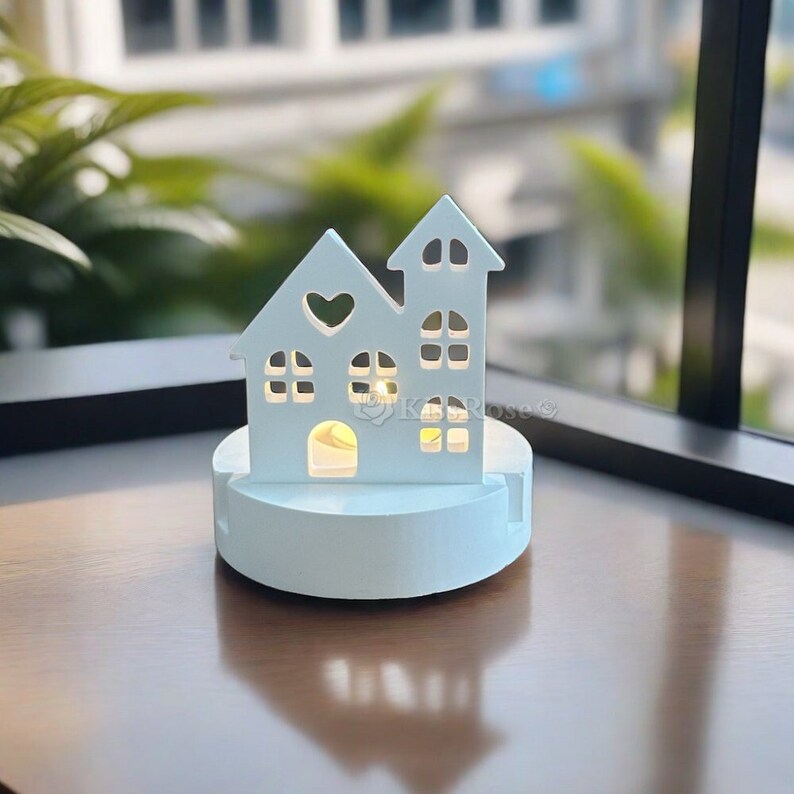 Puede incluir: Portavelas de madera blanca en forma de casa con un peque&ntilde;o coraz&oacute;n recortado en la parte delantera. La casa est&aacute; iluminada desde el interior con una luz blanca c&aacute;lida. El portavelas est&aacute; sobre una superficie de madera.