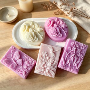Peut inclure: Cinq savons décoratifs avec des motifs floraux et d'insectes en relief. Deux savons ovales crème et rose avec des roses, et trois savons rectangulaires roses avec un papillon, une libellule avec des fleurs et de petites fleurs.