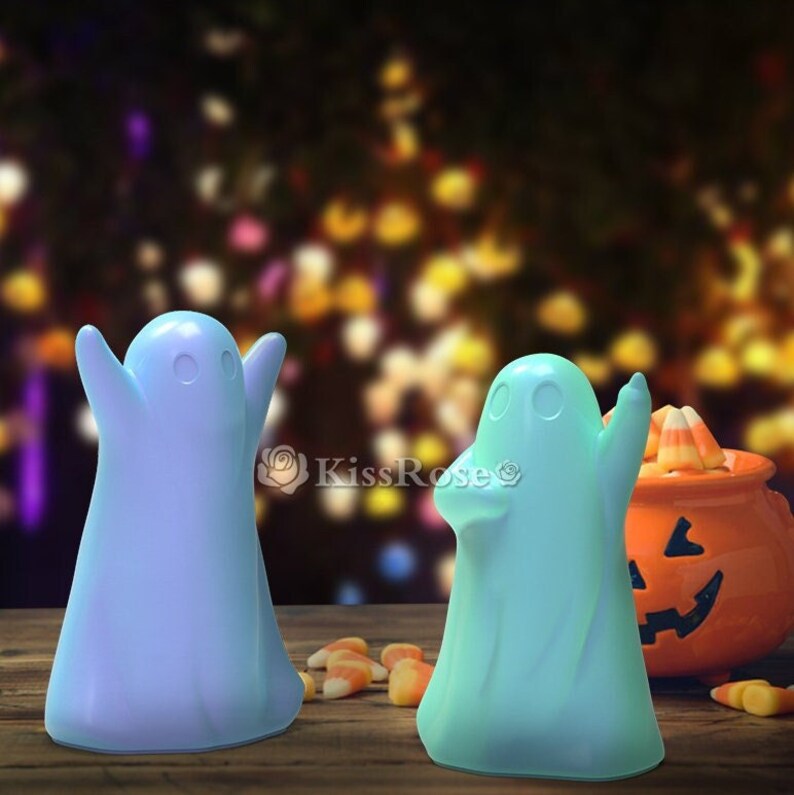 May include: Two ghost-shaped figurines, one light blue and one light green, are standing on a wooden surface. The figurines are in front of a blurred background of candy corn and a jack-o-lantern.