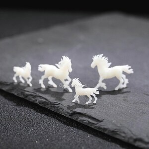 Op de afbeelding: Vier witte plastic paardenfiguurtjes in verschillende maten, gerangschikt in een rij op een zwart leisteen oppervlak.