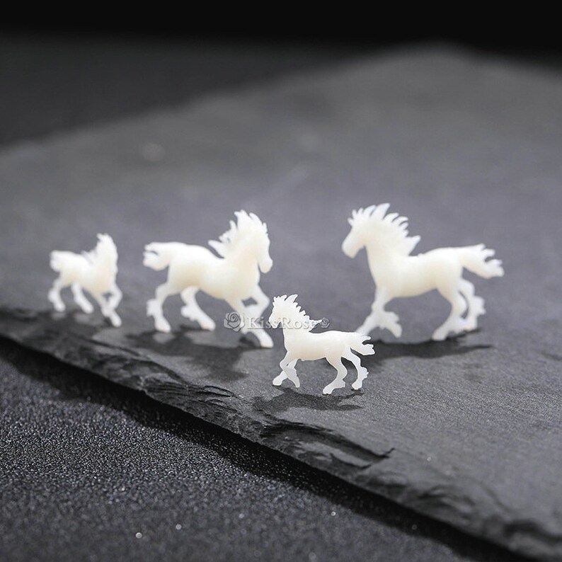 Op de afbeelding: Vier witte plastic paardenfiguurtjes in verschillende maten, gerangschikt in een rij op een zwart leisteen oppervlak.