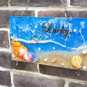 May include: A blue and white resin sign with a mermaid figurine, seashells, and the word "Lucky" in gold lettering. The sign is hanging on a brick wall.