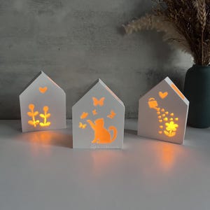 Puede incluir: Tres casas de madera blancas con bombillas en el interior, cada una con un diseño diferente. La primera casa tiene un diseño de flores, la segunda casa tiene un diseño de gato y la tercera casa tiene un diseño de regadera.