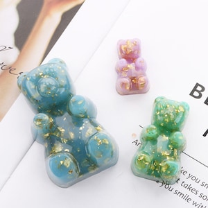May include: Three colorful gummy bear-shaped resin figurines. The largest is teal blue, the smallest is green, and the middle one is purple. All are embedded with gold flakes. The bears are on a white surface.