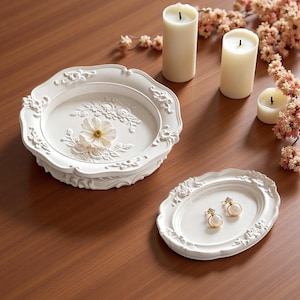 May include: A round white decorative tray with embossed floral details and a single white flower in the centre, alongside a smaller oval white tray holding a pair of gold-toned earrings with white spherical accents, featuring scalloped edges.