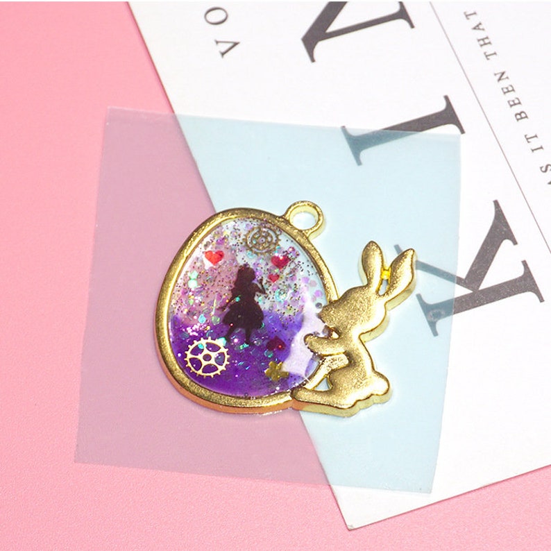 Pode incluir: Um pingente dourado com um camafeu de resina roxa e branca com glitter e uma silhueta de uma menina. O camafeu &eacute; rodeado por uma moldura dourada e uma figura de coelho dourada.