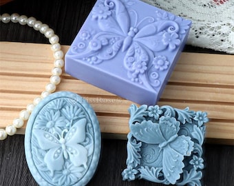 Könnte beinhalten: Drei Silikonformen zum Herstellen von Seife oder Kerzen. Jede Form zeigt ein detailliertes Schmetterlingsdesign mit floralen Akzenten. Die Formen sind in Blautönen und Lila.