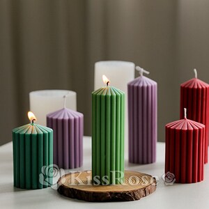 May include: Six pillar candles with a ribbed design in various colors, including green, purple, red, and white. The candles are lit and arranged on a wooden base.