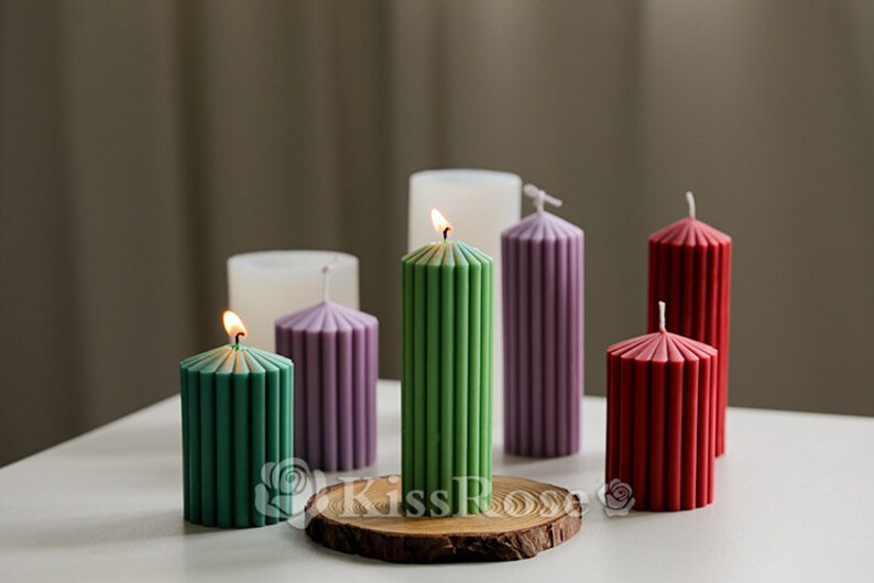 May include: Six pillar candles with a ribbed design in various colors, including green, purple, red, and white. The candles are lit and arranged on a wooden base.
