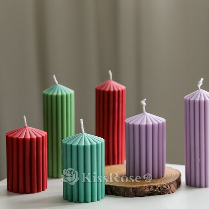 May include: Six pillar candles in various colors, including red, green, and purple. The candles have a fluted design and are arranged on a wooden surface.