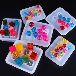 Pode incluir: Seis moldes de silicone para fazer joias de resina. Os moldes são brancos e têm diferentes formas, incluindo quadrados e diamantes. Os moldes estão cheios de contas de resina coloridas.