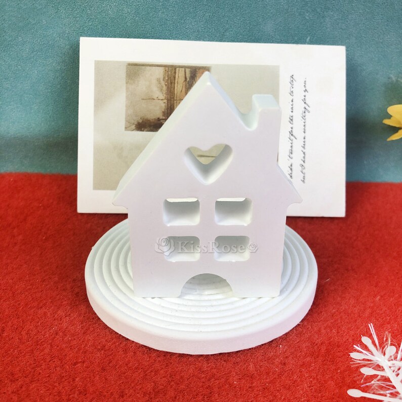 May include: White ceramic house shaped photo holder with a heart cutout and a small chimney. The holder is sitting on a white circular base with concentric rings.