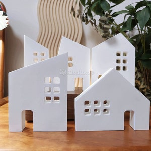 May include: Five white ceramic houses with cut-out windows and doors. The houses are arranged in a row on a wooden surface.
