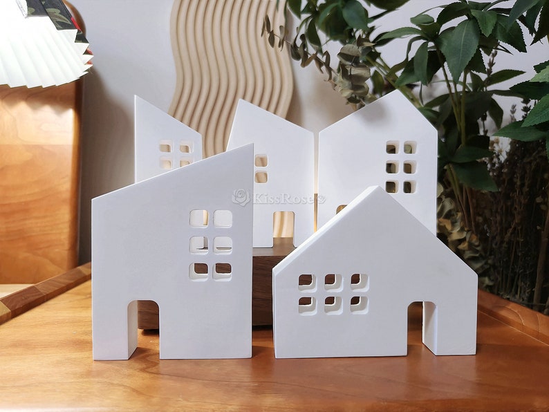 May include: Five white ceramic houses with cut-out windows and doors. The houses are arranged in a row on a wooden surface.
