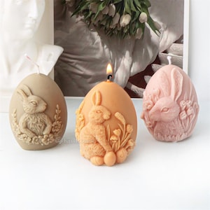May include: Three Easter-themed candles shaped like eggs with bunny rabbit designs. The candles are in shades of brown, pink, and beige.