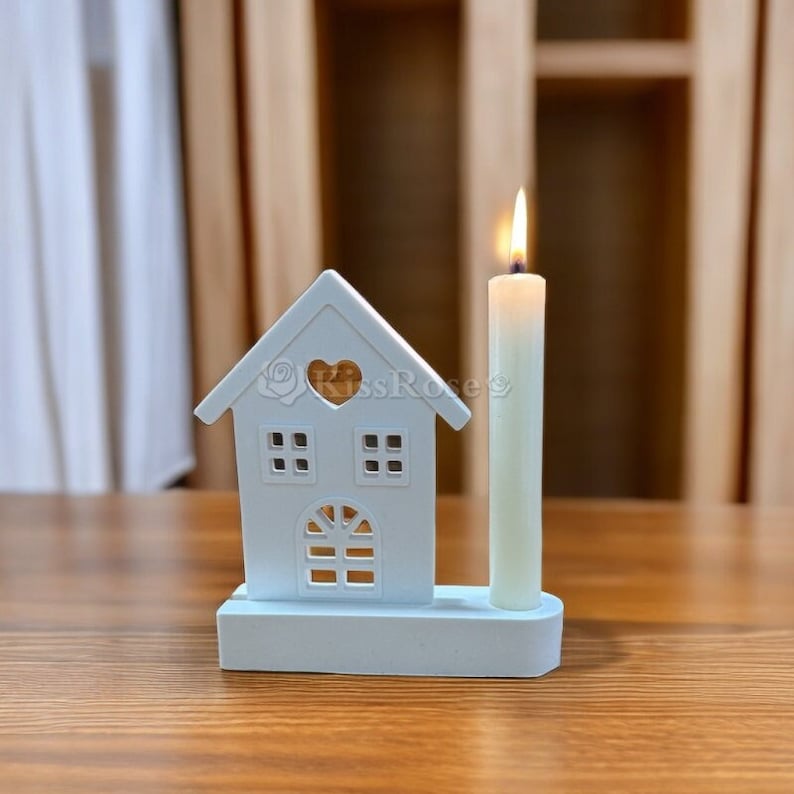 May include: A white wooden house-shaped candle holder with a heart cutout and two windows. A white candle is placed in the holder.
