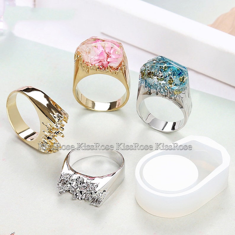 May include: Four resin rings with different colored flowers and gold accents. The rings are on a white surface with a white silicone mold for making rings.