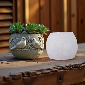 May include: A grey ceramic plant pot with a relief design of birds and flowers, containing green succulents. Next to it, a white, spherical candle holder. The items are on a wooden surface.