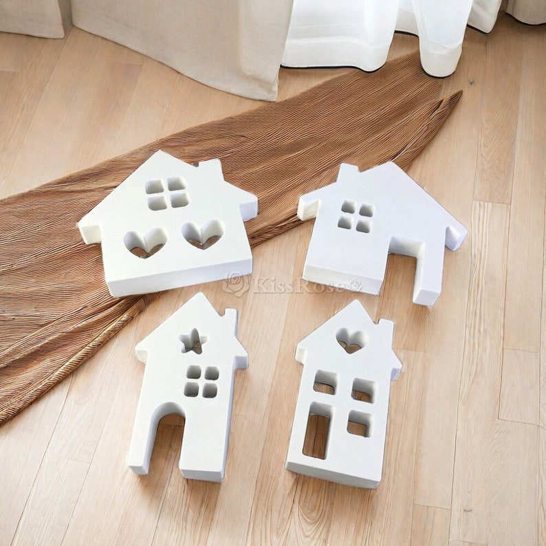 Pu&ograve; includere: Quattro ornamenti in legno bianco a forma di casa con diversi disegni di finestre e porte. Gli ornamenti sono disposti su una superficie marrone.