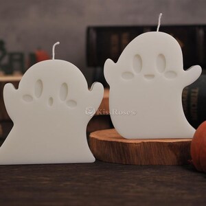 Op de afbeelding: Twee witte kaarsen in de vorm van een spook met schattige gezichtjes. De kaarsen staan op een houten oppervlak.