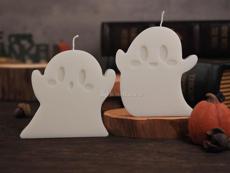 Op de afbeelding: Twee witte kaarsen in de vorm van een spook met schattige gezichtjes. De kaarsen staan op een houten oppervlak.