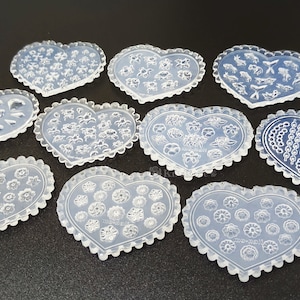 May include: Nine clear heart-shaped silicone molds for nail art. The molds feature various designs, including flowers, chains, and abstract shapes.