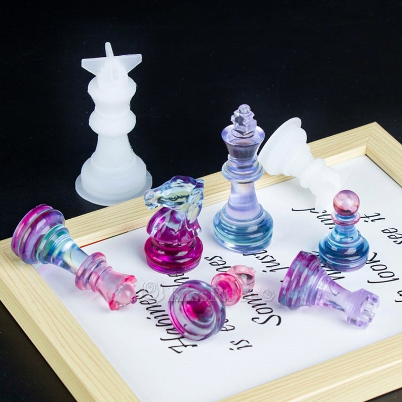 K&ouml;nnte beinhalten: Ein Set aus bunten gl&auml;sernen Schachfiguren, darunter K&ouml;nig, Dame, Springer, Bauer und Turm. Die Figuren sind aus klarem Glas mit schillernden Farben gefertigt und auf einer wei&szlig;en Oberfl&auml;che mit einer handschriftlichen Nachricht angeordnet.