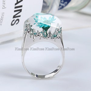 May include: A silver ring with a clear resin setting featuring a teal flower. The ring has a textured band.
