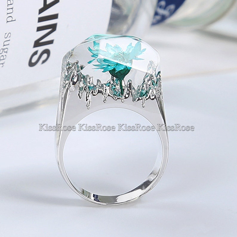 May include: A silver ring with a clear resin setting featuring a teal flower. The ring has a textured band.