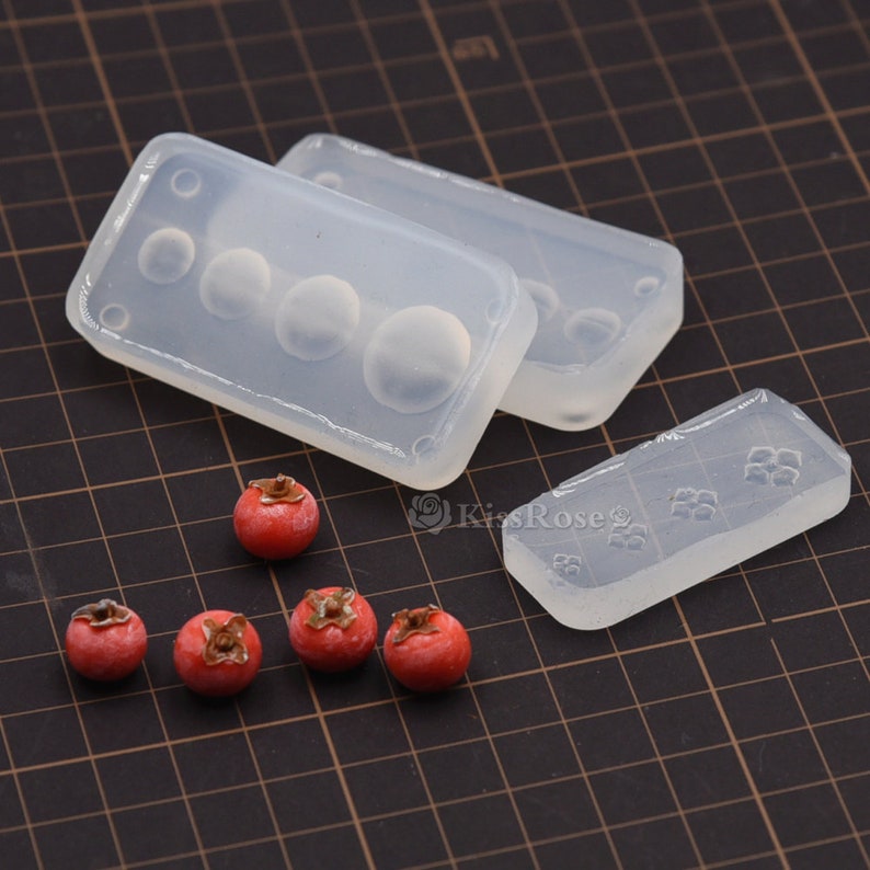 Puede incluir: Cuatro moldes de silicona transparentes para hacer peque&ntilde;os objetos redondos, como fruta en miniatura. Los moldes est&aacute;n dispuestos sobre una superficie de rejilla negra. Seis peque&ntilde;os objetos redondos rojos tambi&eacute;n est&aacute;n sobre la superficie.