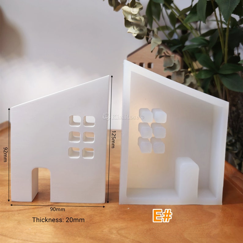 May include: White silicone mould shaped like a house with a door and windows. The mould is 90mm wide, 92mm tall, and 20mm thick. The mould is designed to create a small house-shaped candle or soap.