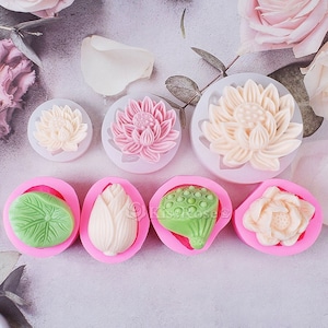 Op de afbeelding: Een set van zes roze siliconen mallen voor het maken van lotusbloem- en bladvormen. De mallen zijn gevuld met witte en roze lotusbloemen, een groen blad en een witte knop.