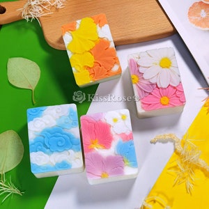 May include: Four rectangular bars of soap with different floral and geometric designs. The soaps are white with various colors, including blue, pink, orange, and yellow.