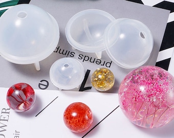 May include: A set of clear silicone molds for making round resin ornaments. The molds are shown with several finished resin ornaments, including a gold glitter ornament, a red and white marbled ornament, and a pink ornament with dried flowers.